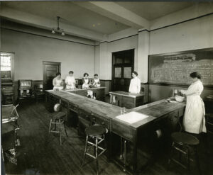 Women stand around large table in domestic science classroom at the Alberta Ladies College.